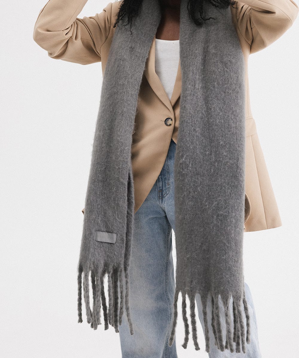 A person wearing a beige blazer and blue jeans, and a grey oversized knit scarf with fringes, draped over the neck against a plain background  #color_grey