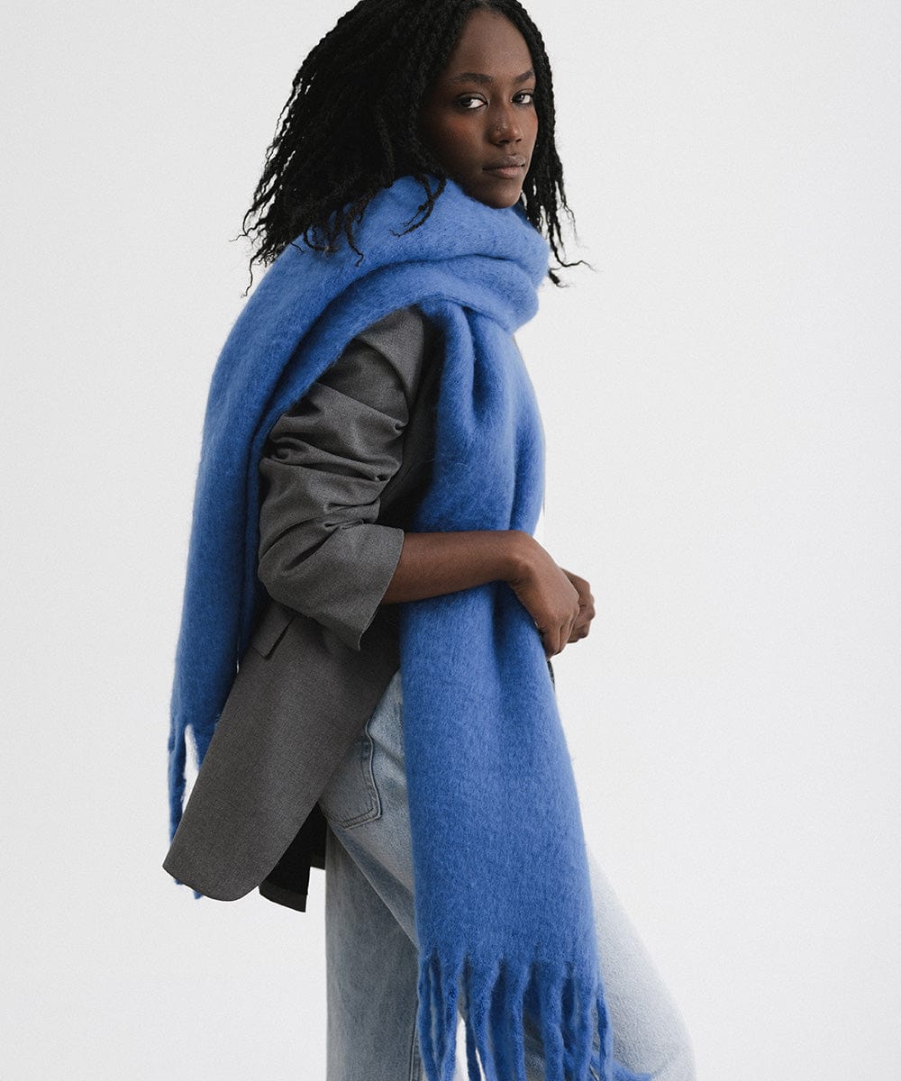 A woman wearing a grey blazer and blue jeans, and an alpine blue colored oversized knit scarf with fringes, draped over the neck against a plain background  #color_alpine blue