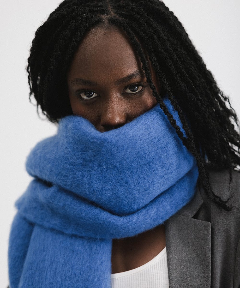 A person wearing an alpine blue colored oversized knit scarf with fringes, draped over the neck against a plain background  #color_alpine blue