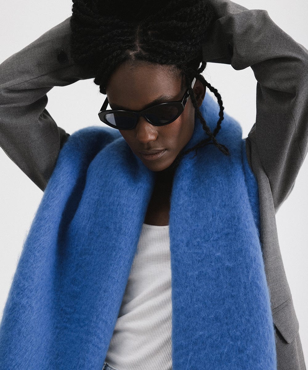 A woman wearing sunglasses, a grey blazer, blue jeans, and an alpine blue colored oversized knit scarf with fringes, draped over the neck against a plain background  #color_alpine blue