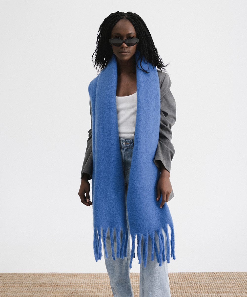 A woman wearing sunglasses, a grey blazer, blue jeans, and an alpine blue colored oversized knit scarf with fringes, draped over the neck against a plain background  #color_alpine blue