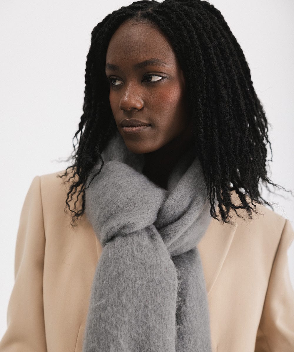 A woman wearing a beige blazer, and a grey oversized knit scarf with fringes, draped over the neck against a plain background  #color_grey