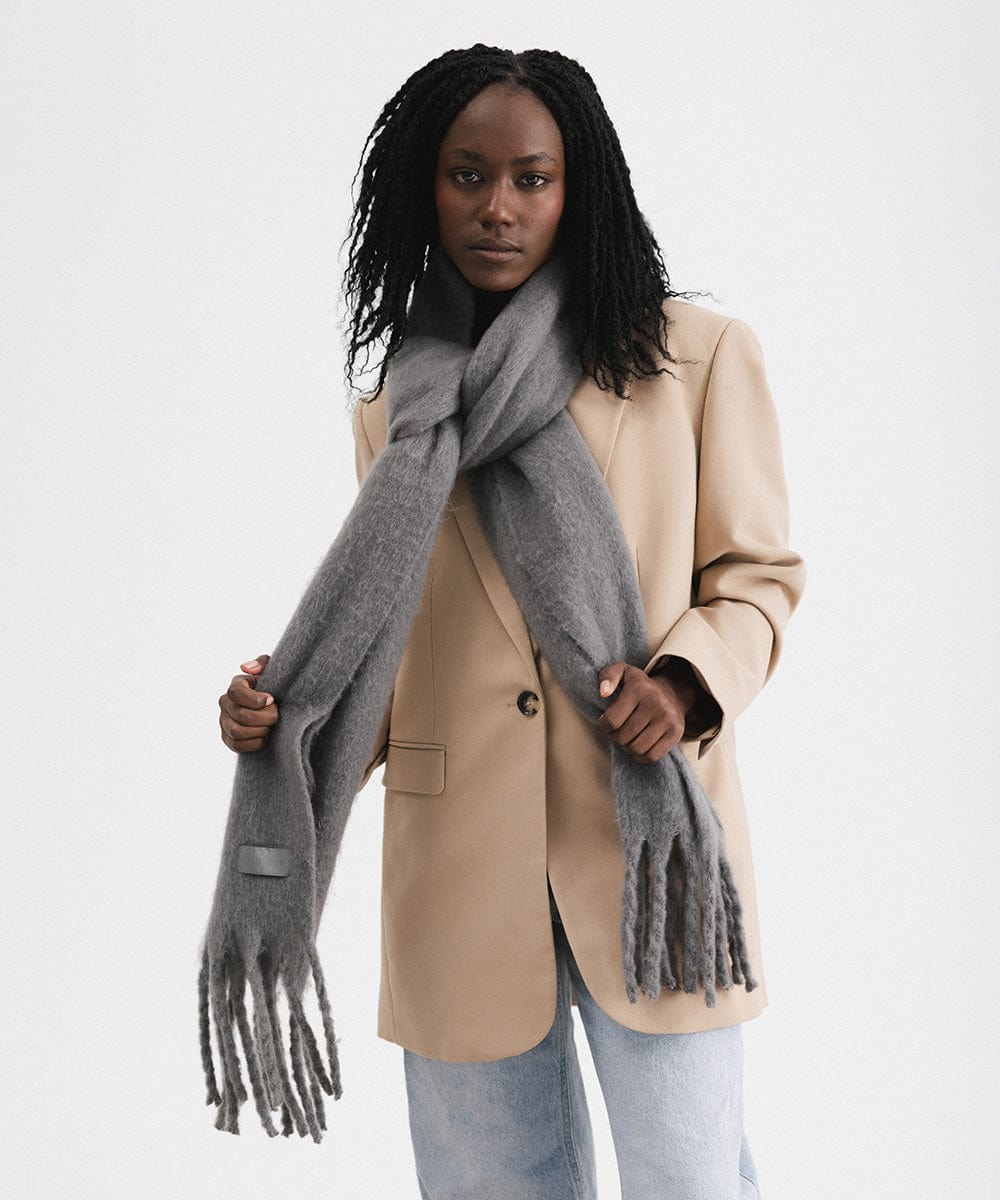A woman wearing a beige blazer and blue jeans, and a grey oversized knit scarf with fringes, draped over the neck against a plain background  #color_grey