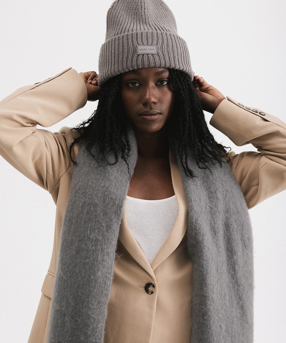 A person wearing a beige blazer and grey beanie, and a grey oversized knit scarf with fringes, draped over the neck against a plain background  #color_grey