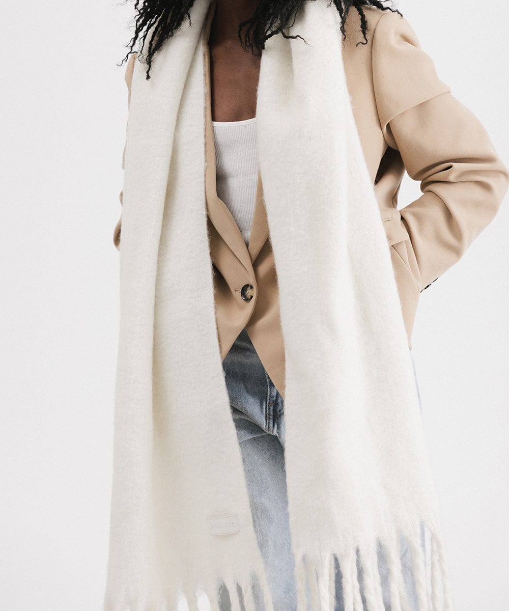 A person wearing a beige blazer, blue jeans, and an off white oversized knit scarf with fringes, draped over the neck against a plain background  #color_off white