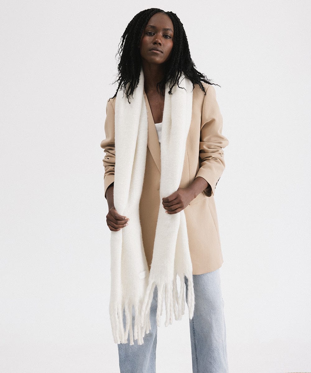 A woman wearing a beige blazer, blue jeans, and an off white oversized knit scarf with fringes, draped over the neck against a plain background  #color_off white