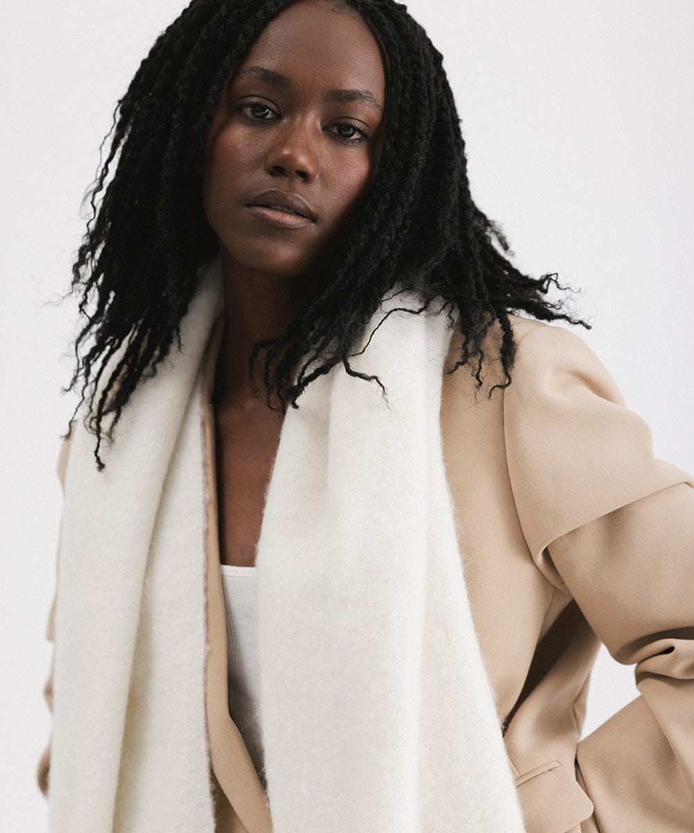 A person wearing a beige blazer and an off white oversized knit scarf with fringes, draped over the neck against a plain background  #color_off white