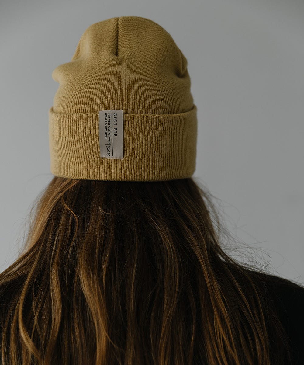 Woman wearing a honeycomb colored beanie with the brand label  and a black coat, facing behind against a plain background #color_honeycomb