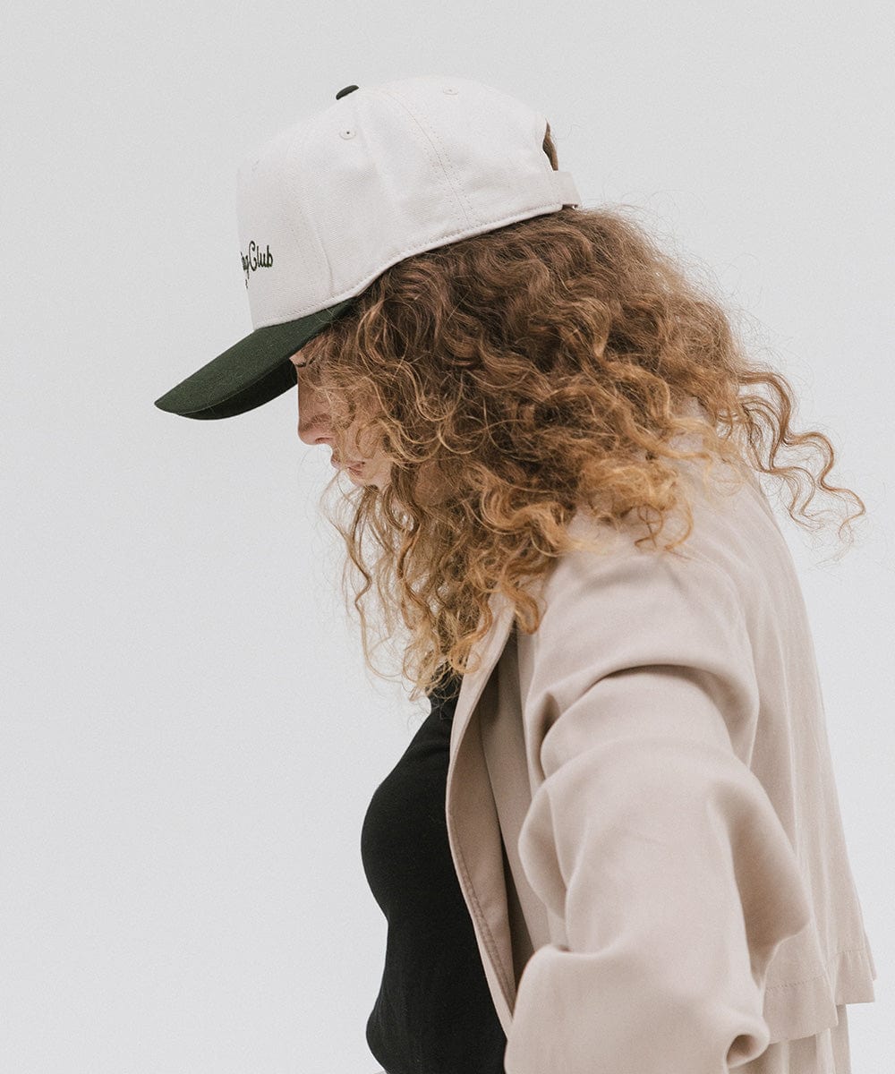 Woman wearing a cream and dark green cap with 'Slow Morning Club' text and a black top with a beige coat, facing sideways against a plain background  #color_cream - dark green