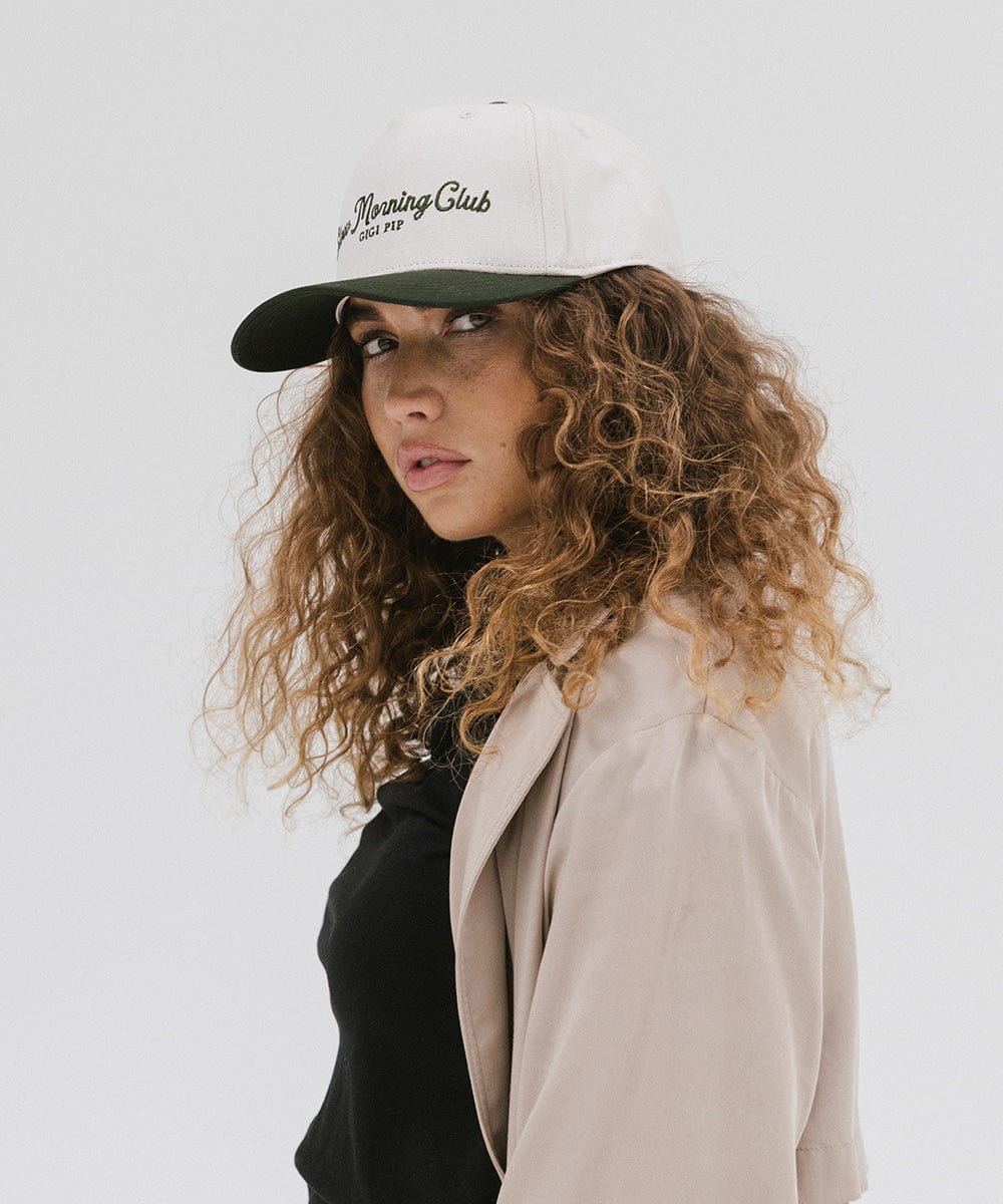Woman wearing a cream and dark green cap with 'Slow Morning Club' text and a black top with a beige coat against a plain background  #color_cream - dark green