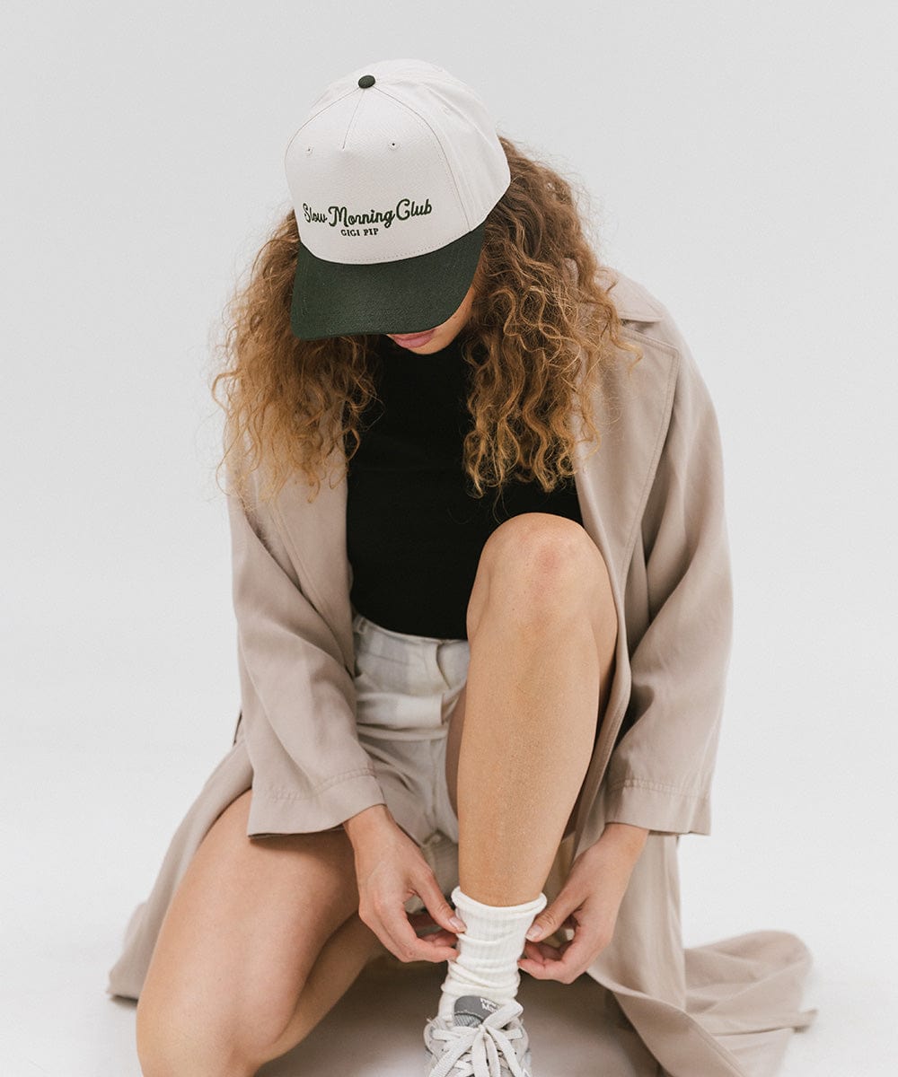Woman wearing a cream and dark green cap with 'Slow Morning Club' text, a black top with a beige coat, and white shorts, sitting against a plain background  #color_cream - dark green