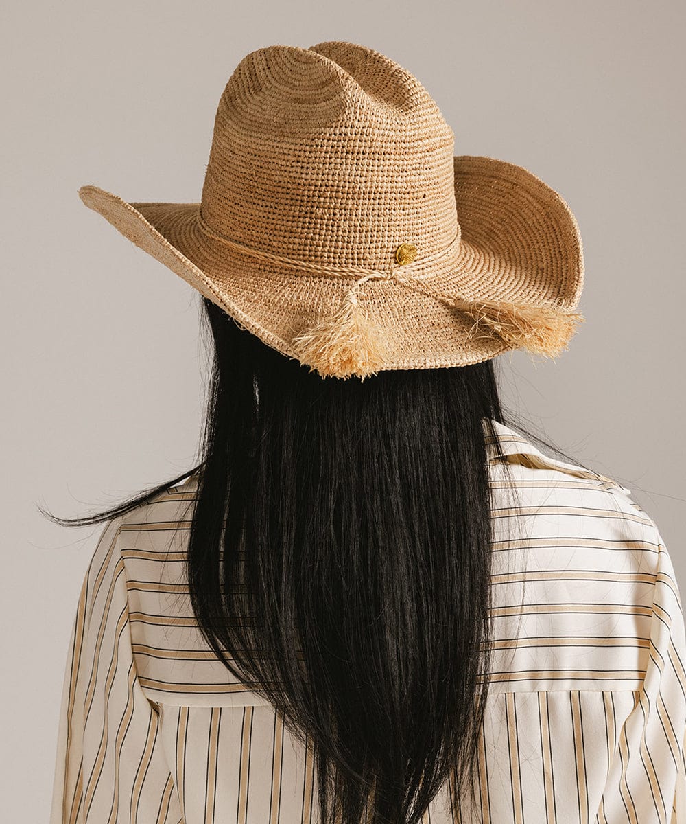 Woman wearing a natural colored straw cowboy hat with a band and a light colored shirt, facing behind against a plain background #color_natural