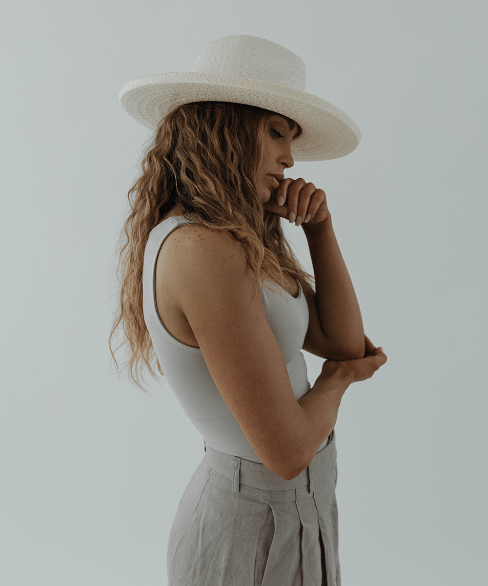Woman wearing a white pencil brim straw hat, a white top and grey pants, posing sideways against a plain background #color_white