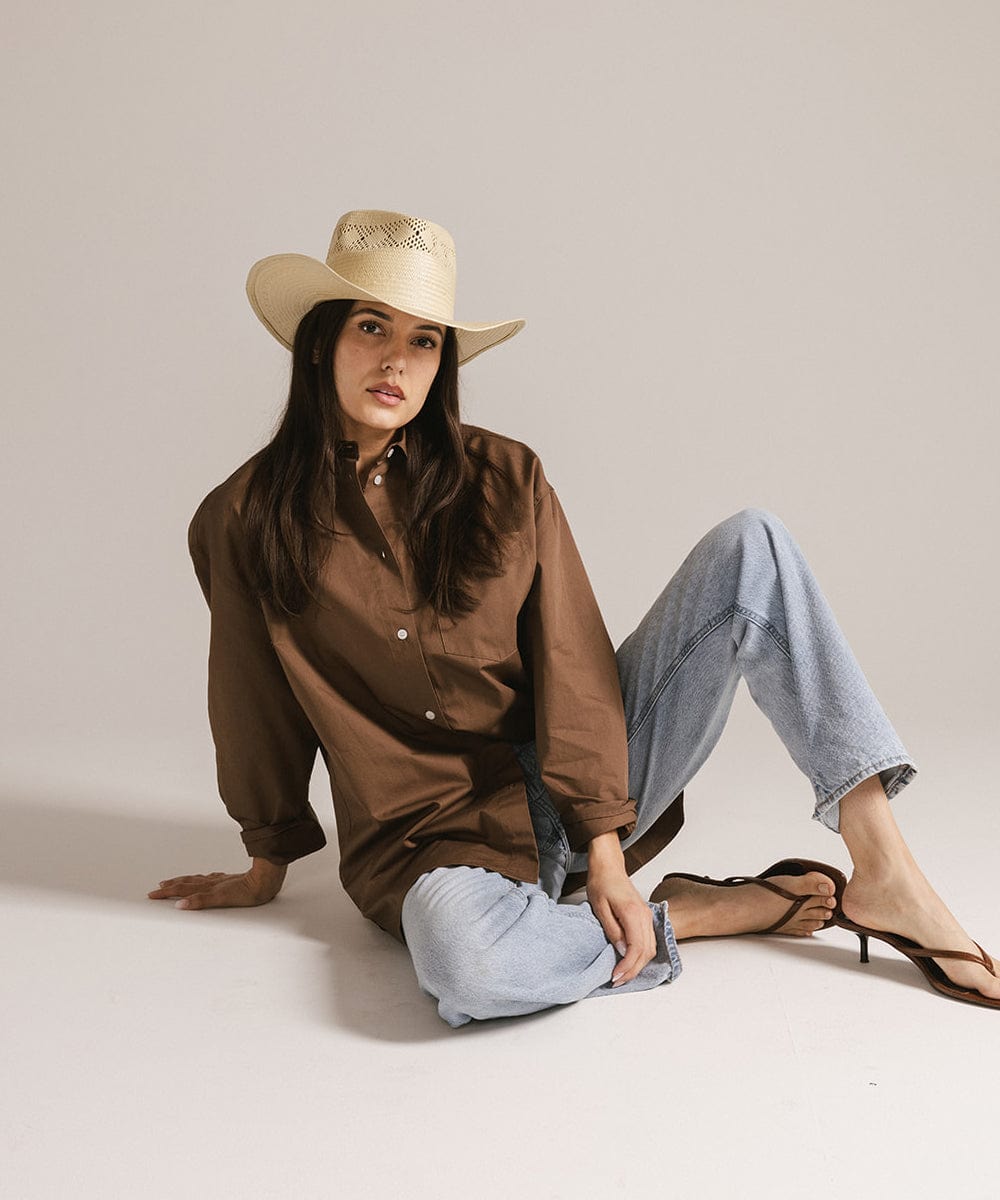 Woman wearing a natural colored straw hat, a brown shirt, blue jeans and brown sandals, sitting against a plain background #color_natural 