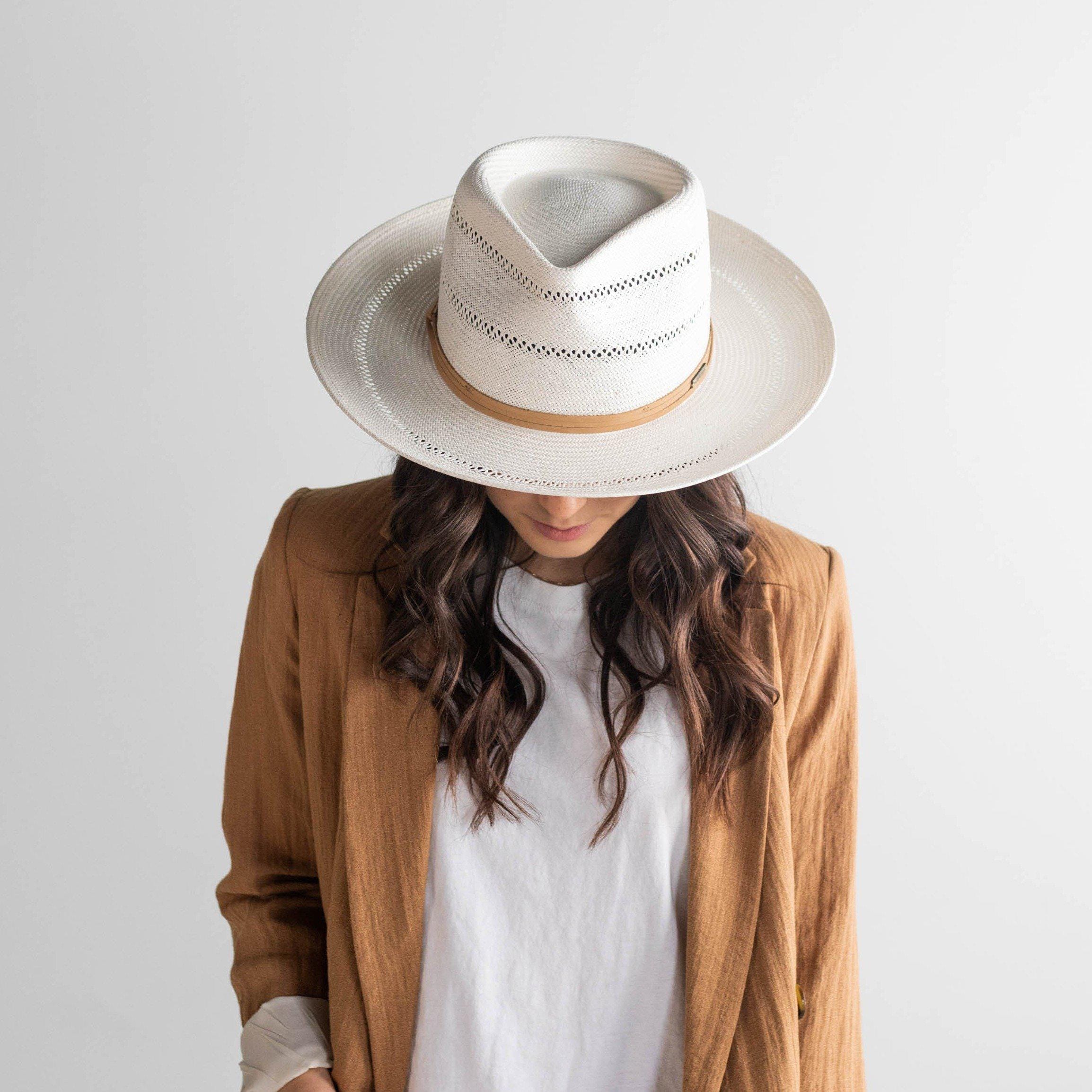 Woman wearing a tan straw fedora hat with a hat band and a white top with a brown coat against a plain background #color_tan