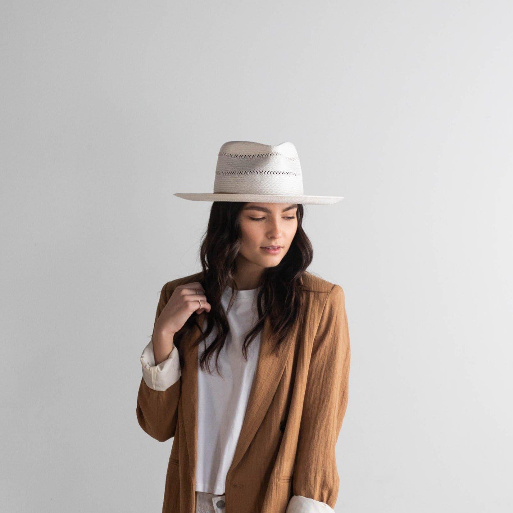Woman wearing a tan straw fedora hat with a hat band and a white top with a brown coat against a plain background #color_tan