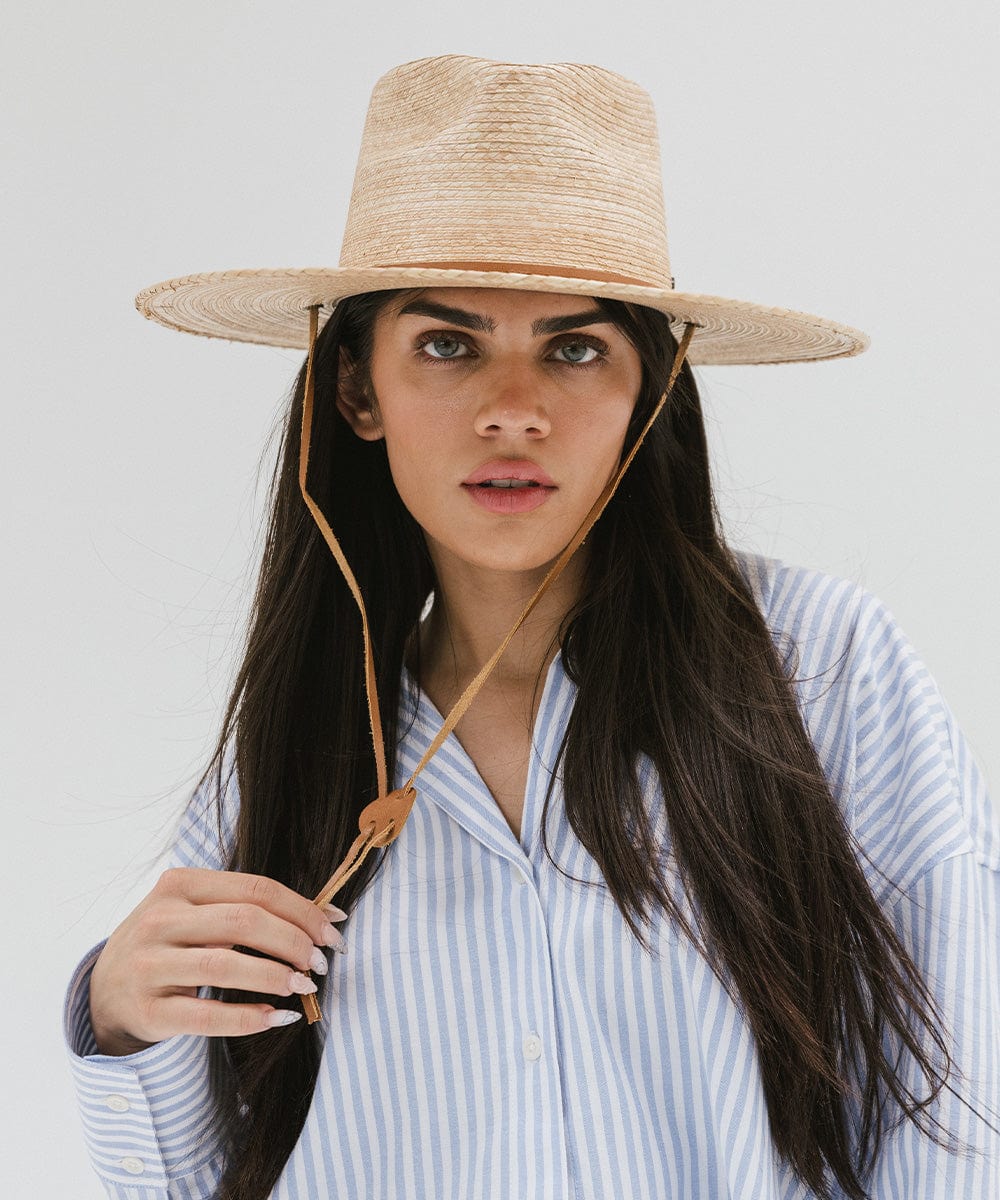 Woman with long hair wearing an oak colored palm straw sun hat with a brown chinstrap and a white shirt with blue lines against a plain background #color_oak