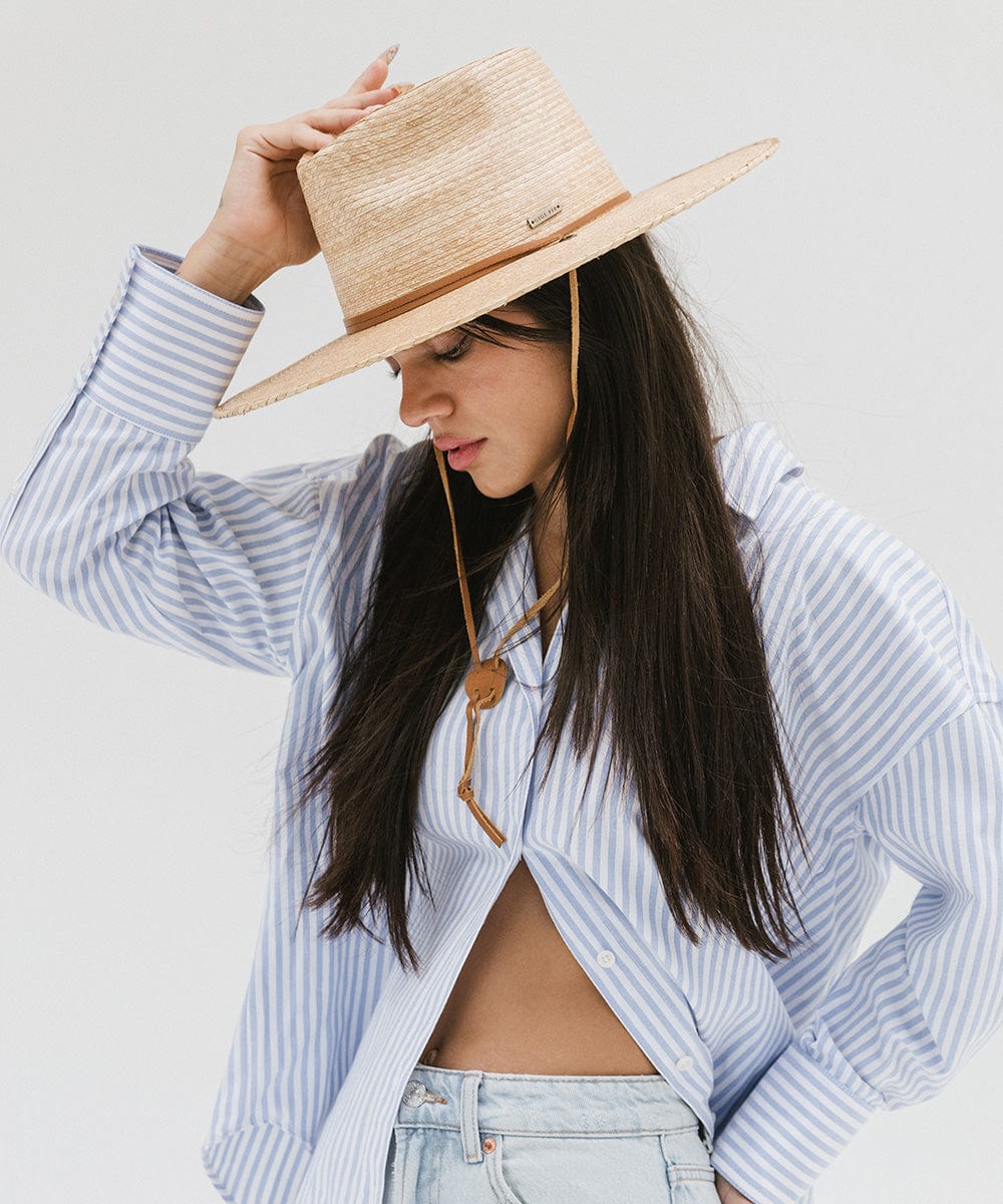 Woman with long hair wearing an oak colored palm straw sun hat with a brown chinstrap and a white shirt with blue lines, posing against a plain background #color_oak
