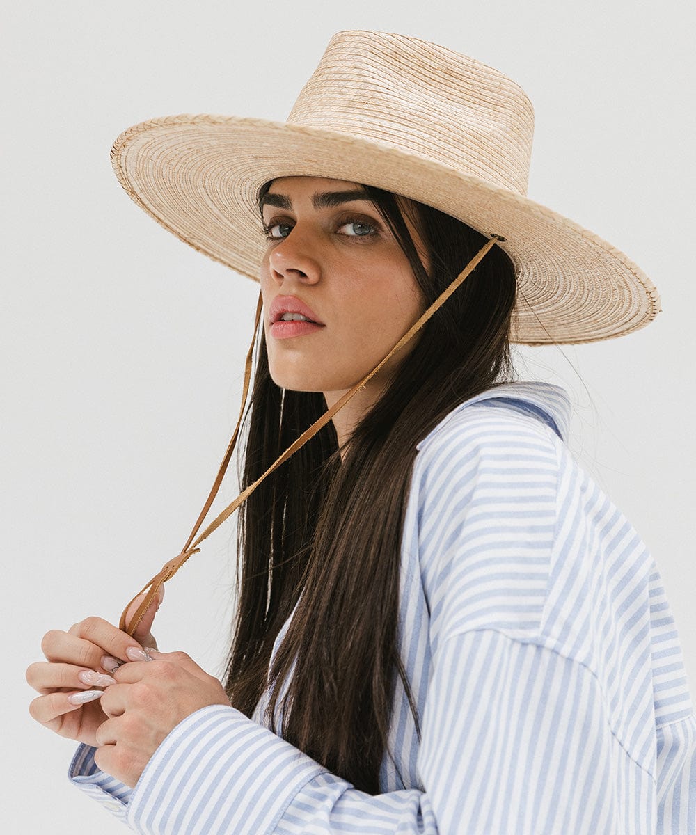 Woman wearing an oak colored palm straw sun hat with a brown chinstrap and a white shirt with blue lines, looking sideways against a plain background #color_oak
