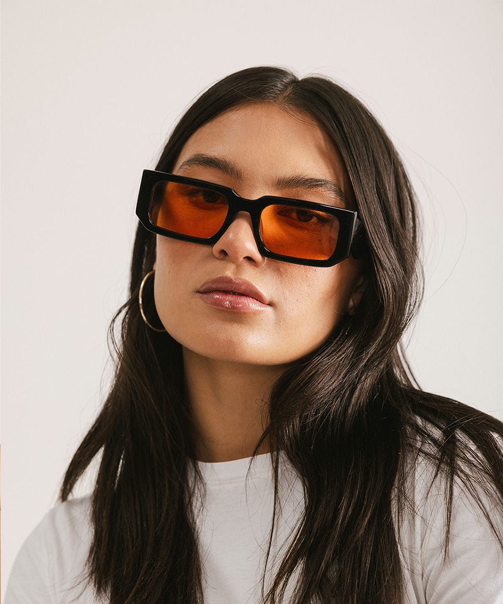Woman wearing sunglasses with black frame and orange lenses and a white t-shirt against a plain background #color_black