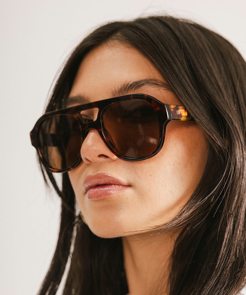 Woman wearing tortoise colored aviator sunglasses against a plain  background #color_tortoise