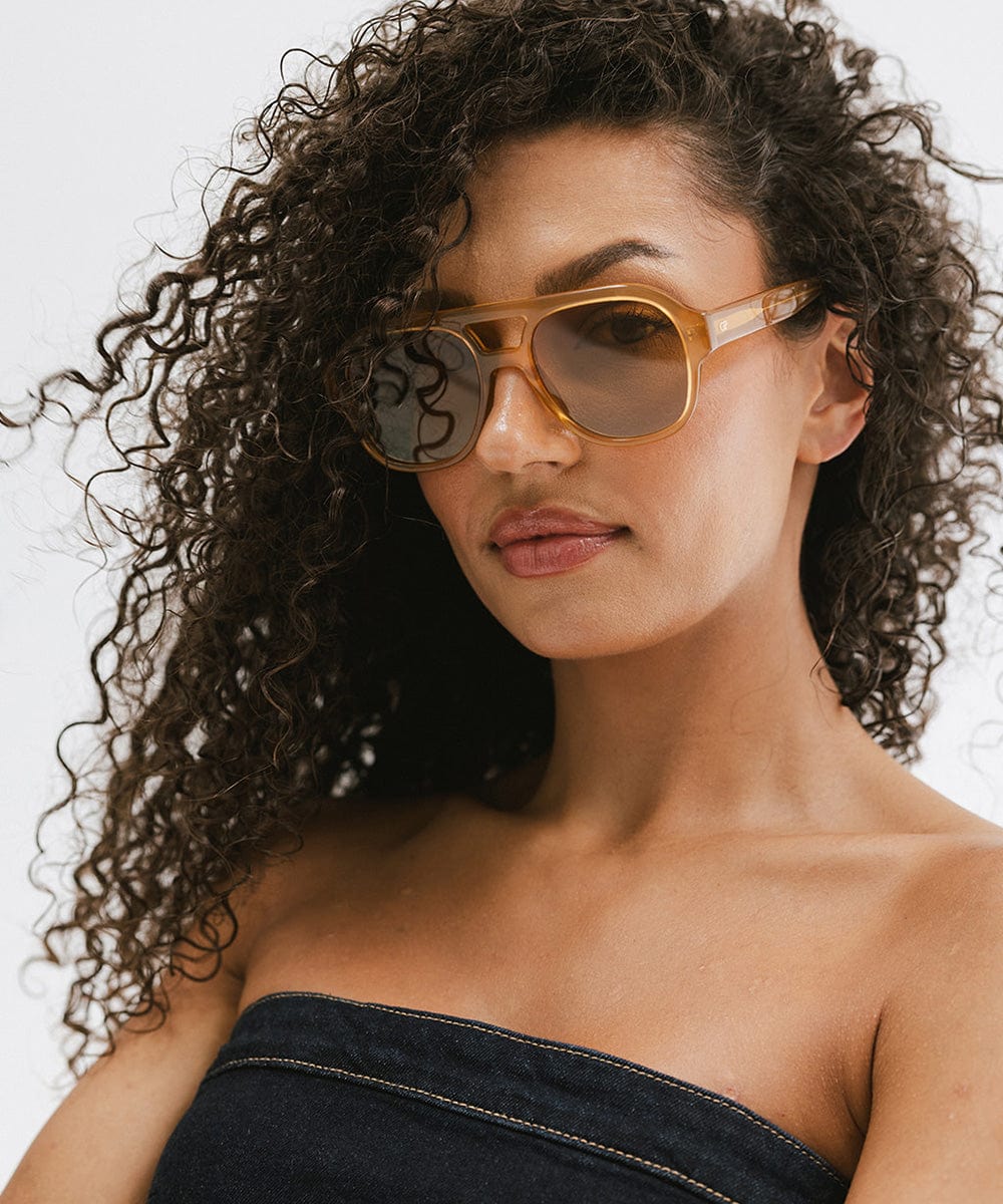 Woman with curly hair wearing honey colored aviator sunglasses and a denim top  against a plain background #color_honey