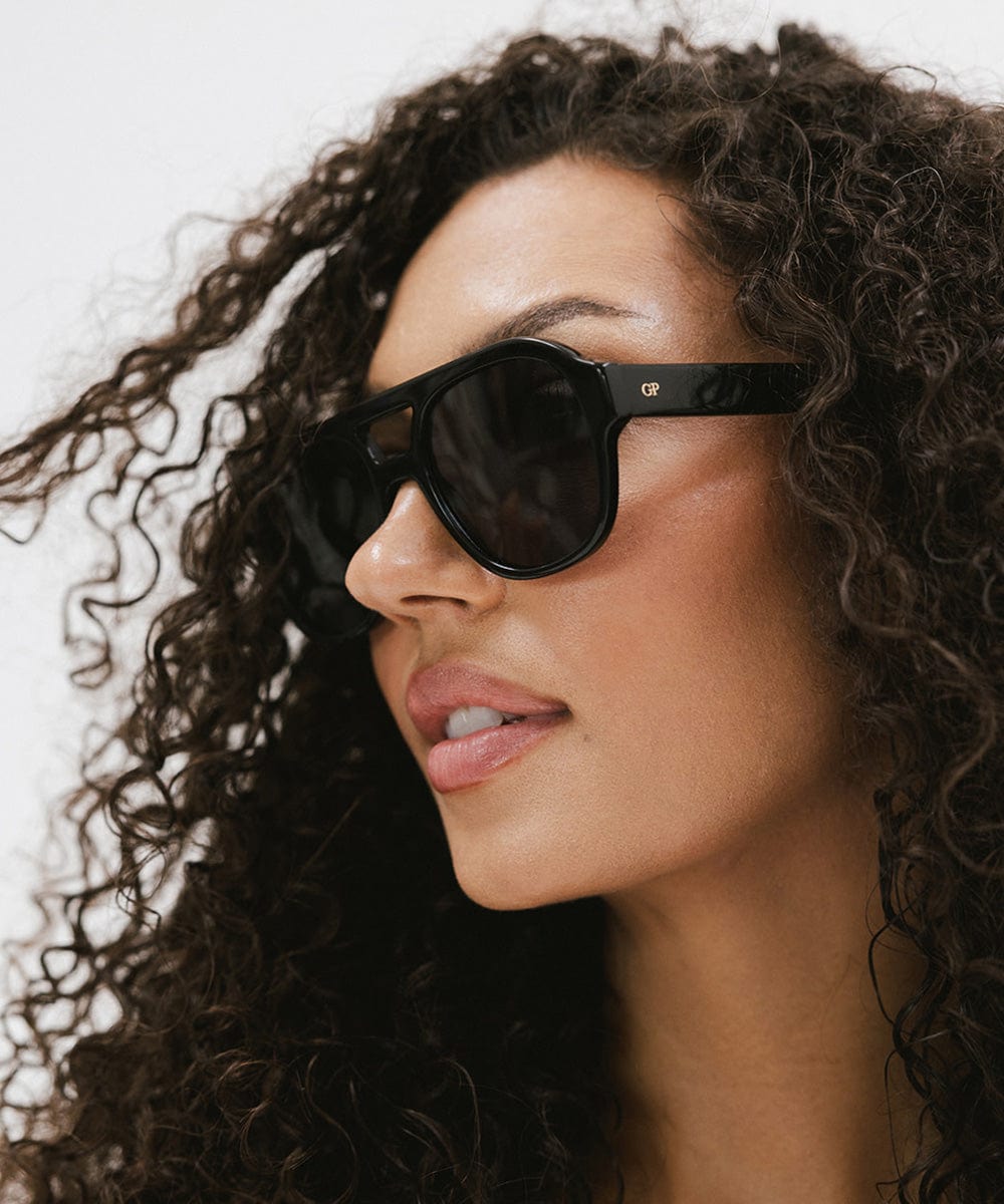 Woman with curly hair wearing black aviator sunglasses against a plain background  #color_black