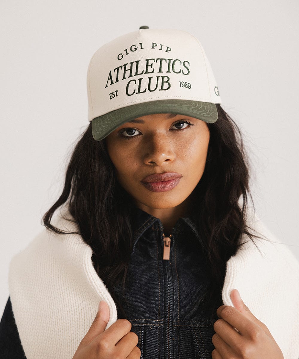 Woman wearing a cream and dark green Trucker hat with 'Gigi Pip Athletics Club' text and a denim jacket against a plain background  #color_cream - dark green