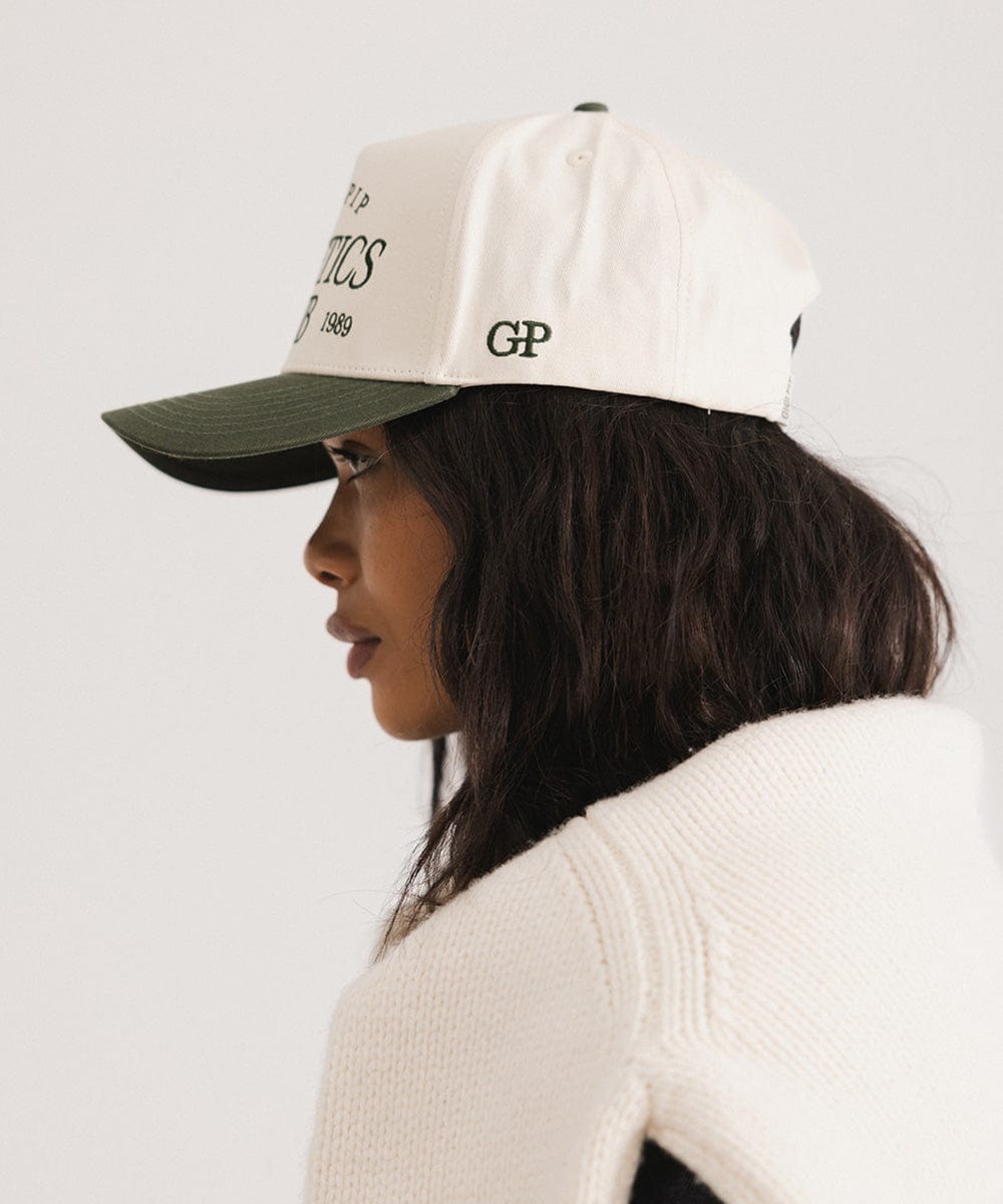 Woman wearing a cream and dark green Trucker hat and a white sweater, facing sideways against a plain background  #color_cream - dark green