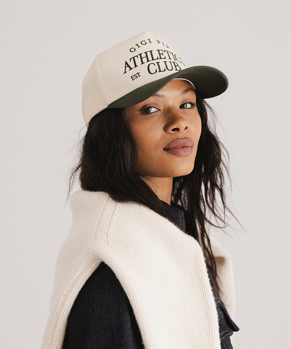 Woman wearing a cream and dark green Trucker hat with 'Gigi Pip Athletics Club' text and a denim jacket, posing sideways against a plain background  #color_cream - dark green