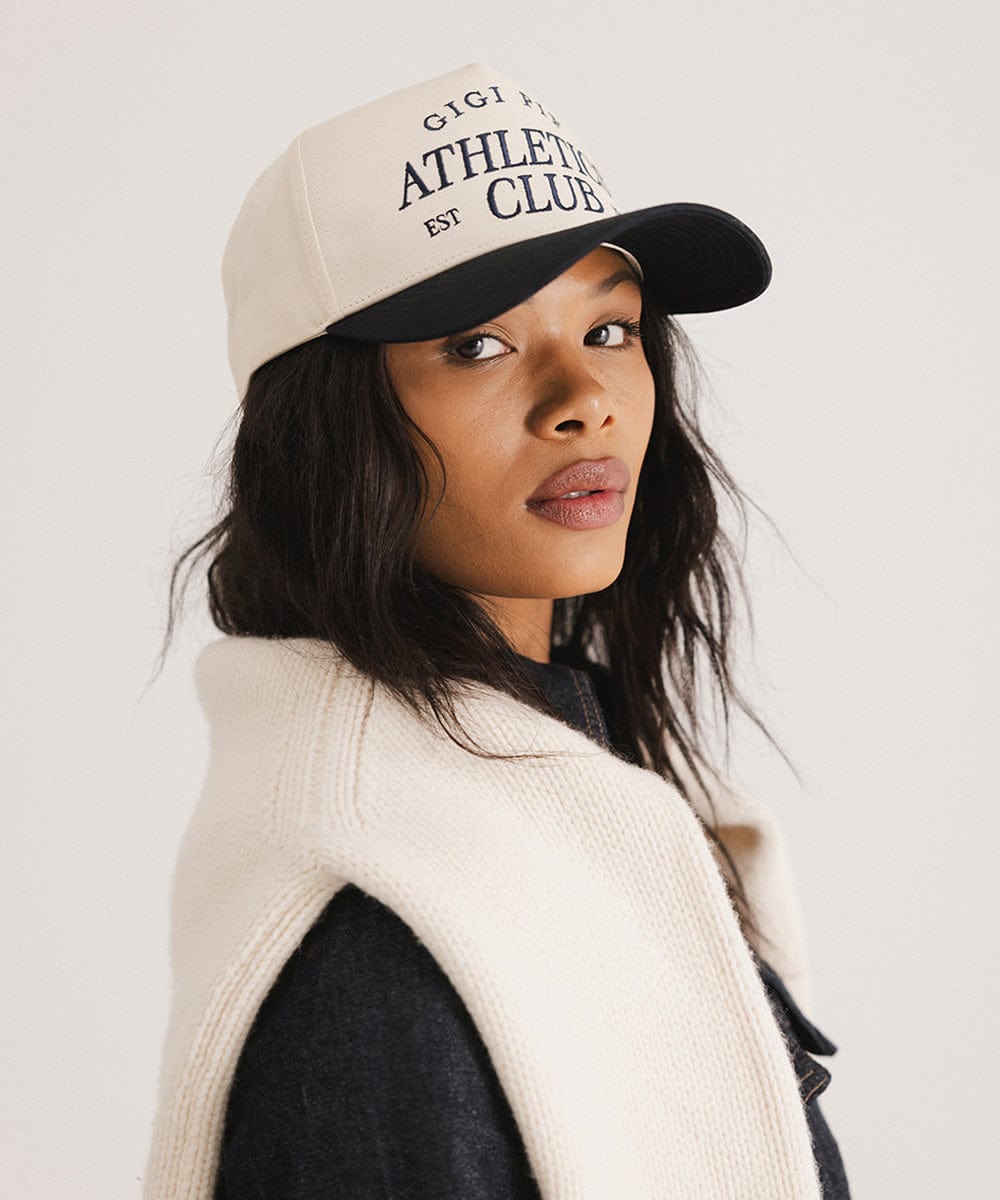 Woman wearing a Navy Trucker hat with 'Gigi Pip Athletics Club' text and a denim jacket, posing sideways  against a plain background #color_navy