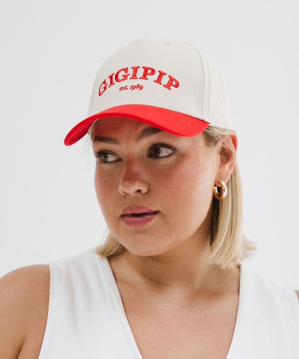 Woman wearing a cream and red trucker hat with 'GIGI PIP' embroidered in red on the front and a white top against a plain background  #color_cream - red