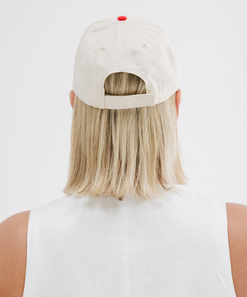 Woman wearing a cream and red trucker hat and a white top against a plain background  #color_cream - red
