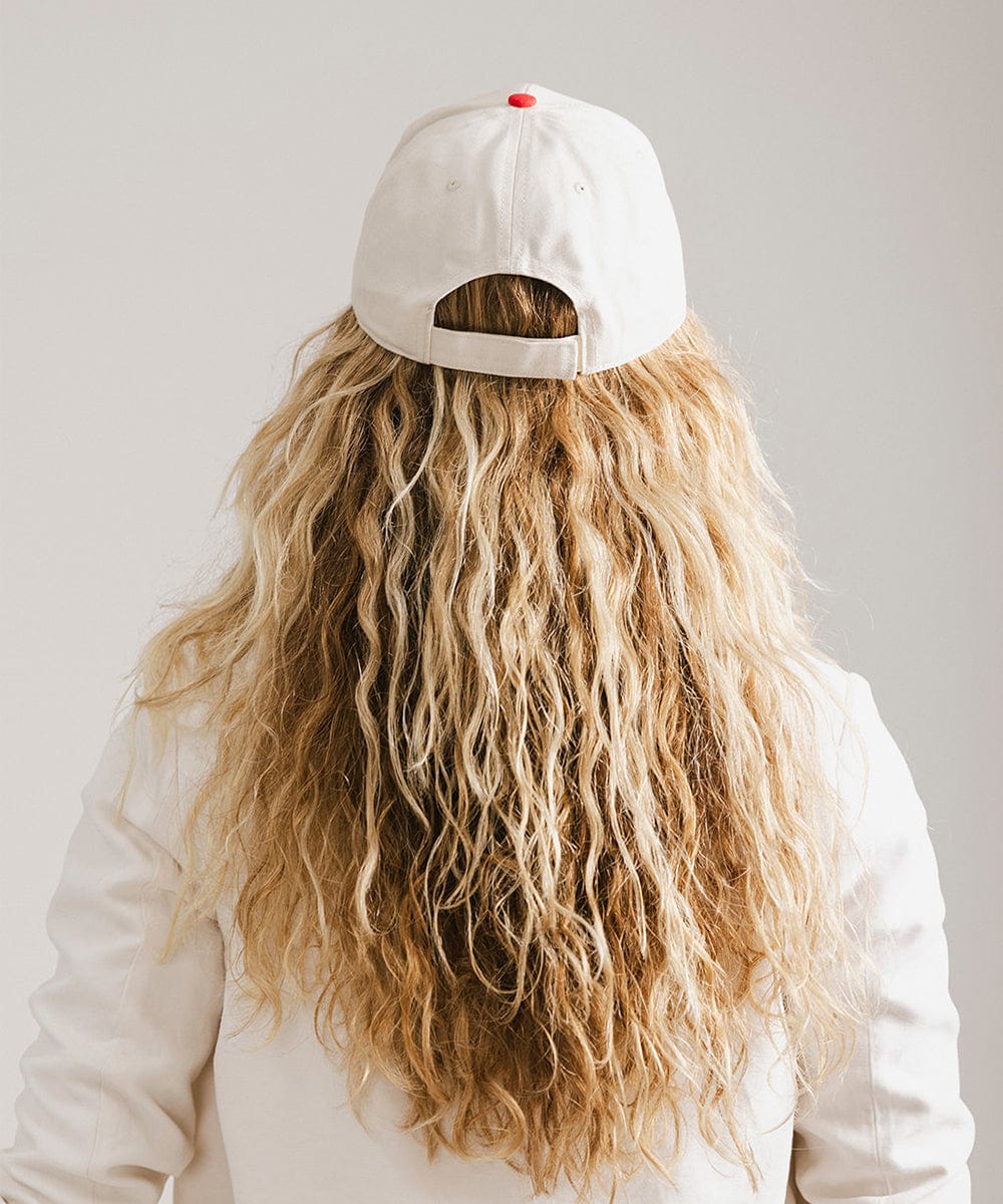 Woman with long hair wearing a cream and red trucker hat and a white coat against a plain background  #color_cream - red