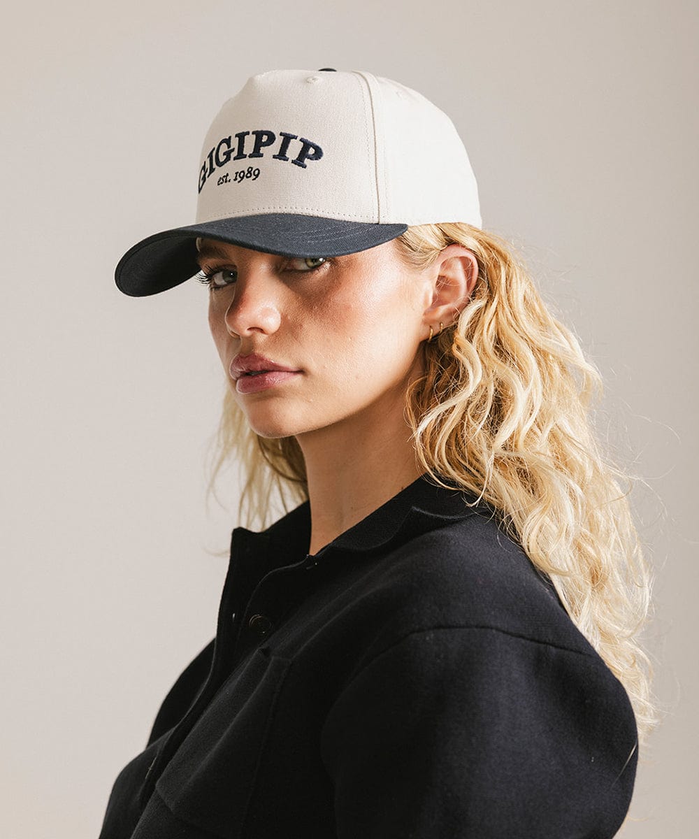 Woman wearing a cream and navy trucker hat with 'GIGI PIP' embroidered in navy on the front and  a black shirt, posing sideways  against a plain background  #color_cream - navy
