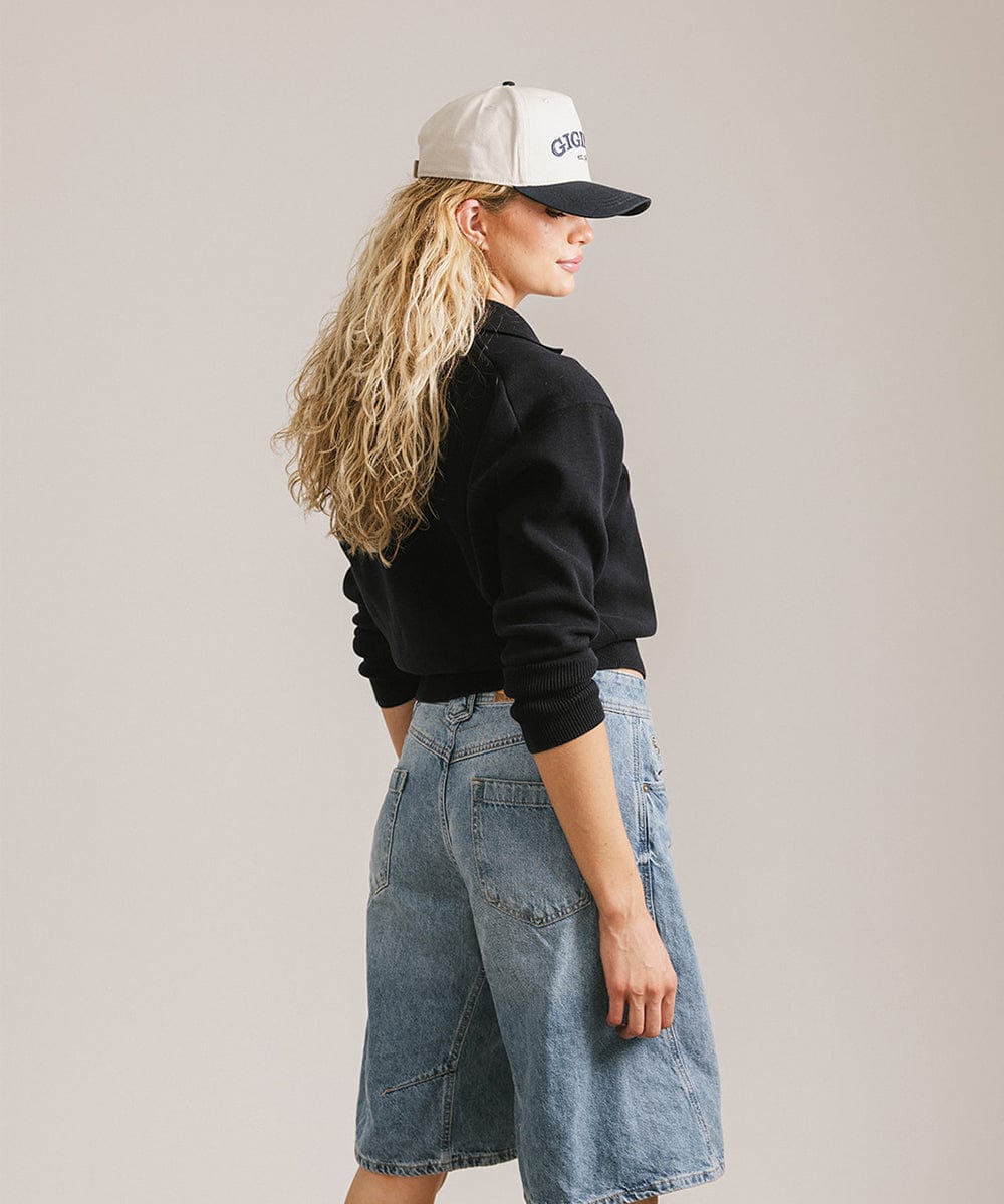Woman wearing a cream and navy trucker hat, a black sweater and denim shorts, posing sideways against a plain background  #color_cream - navy