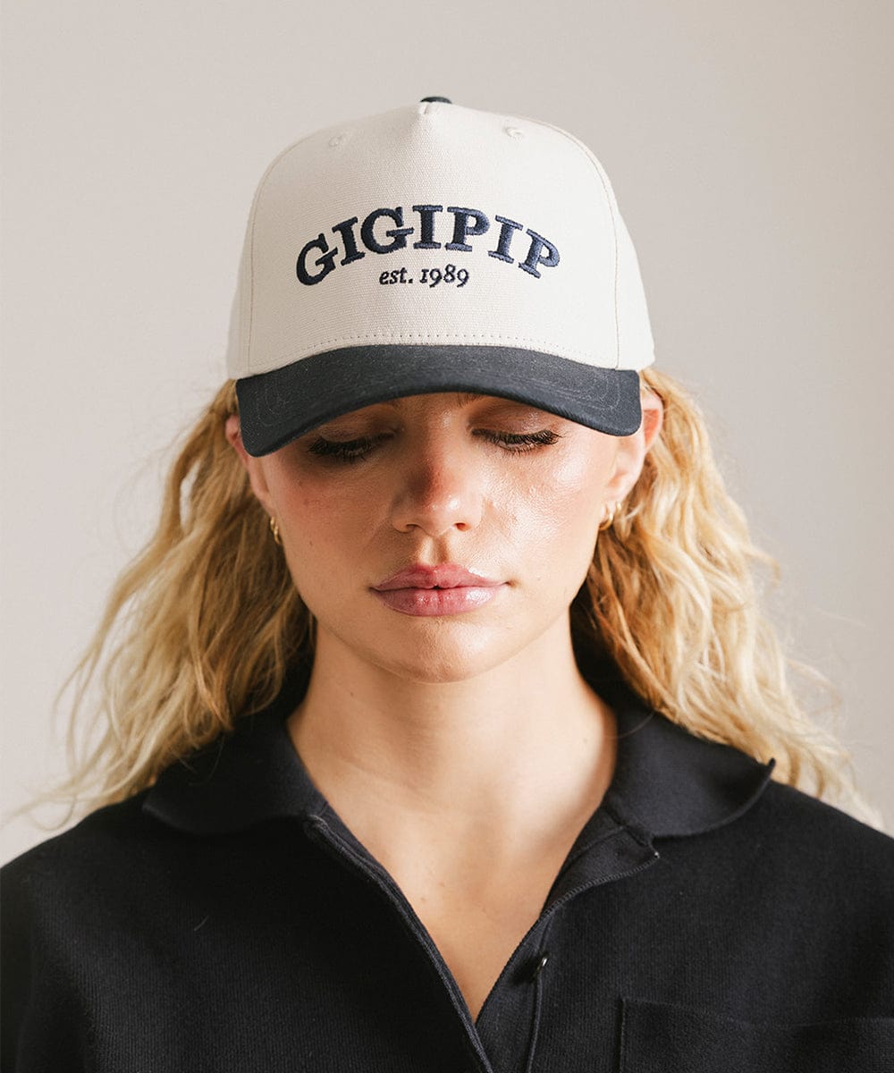 Woman wearing a cream and navy trucker hat with 'GIGI PIP' embroidered in navy on the front and  a black shirt against a plain background  #color_cream - navy