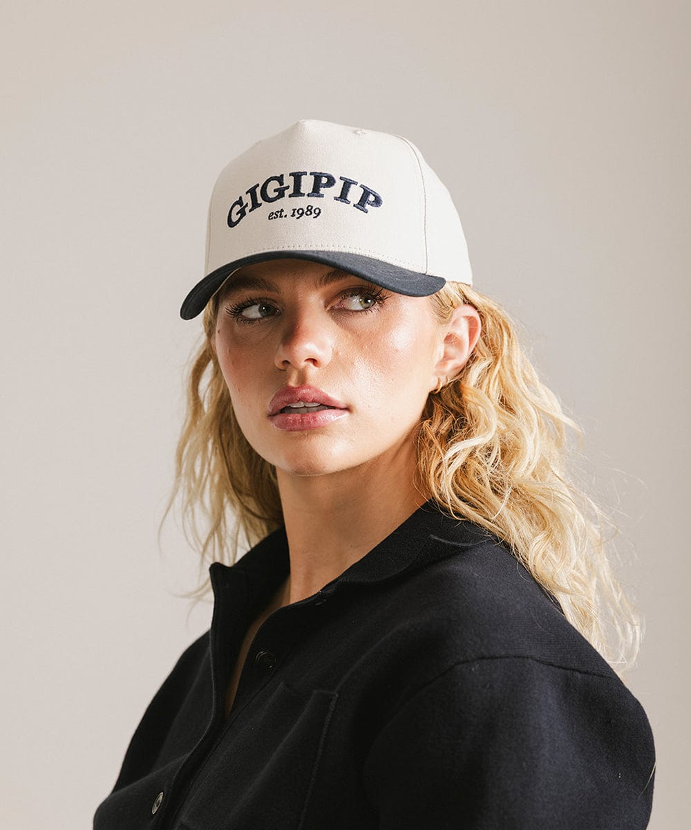 Woman wearing a cream and navy trucker hat with 'GIGI PIP' embroidered in navy on the front and  a black shirt, posing against a plain background  #color_cream - navy