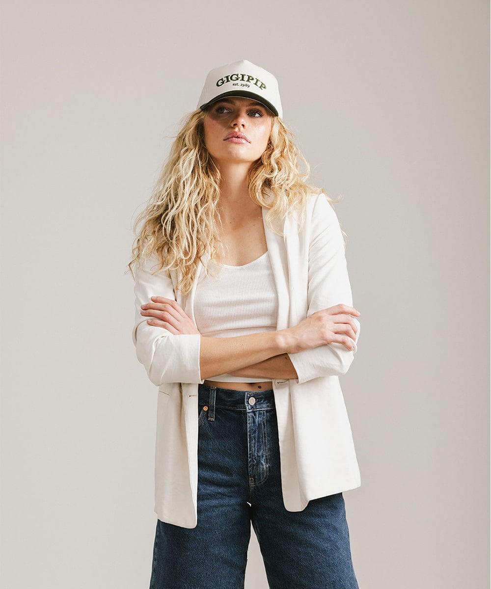 Woman wearing a cream and dark green trucker hat with 'GIGI PIP' embroidered in dark green on the front, a white top with a white coat and blue jeans  against a plain background #color_cream - dark green