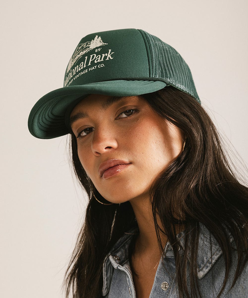 Woman wearing a forest green colored trucker hat with 'National Park' logo against a plain background  #color_forest green