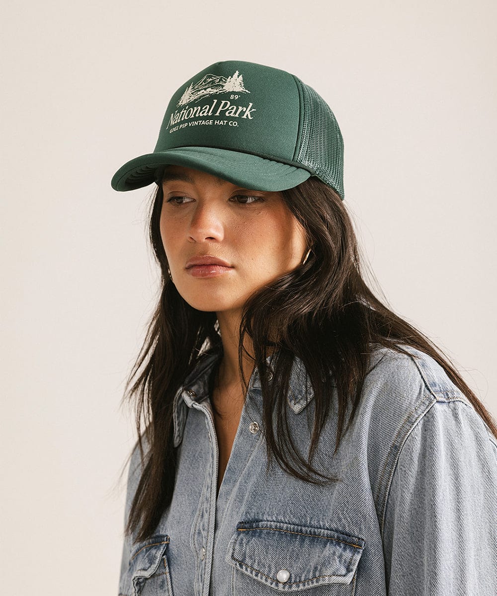 Woman wearing a forest green colored trucker hat with 'National Park' logo and a denim shirt against a plain background  #color_forest green
