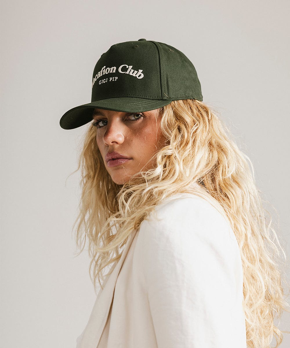 Woman wearing a dark green cap with 'Vacation Club' text and a white coat, posing sideways against a plain background  #color_dark green