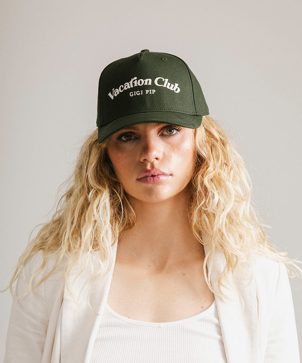 Woman wearing a dark green cap with 'Vacation Club' text and a white coat against a plain background  #color_dark green