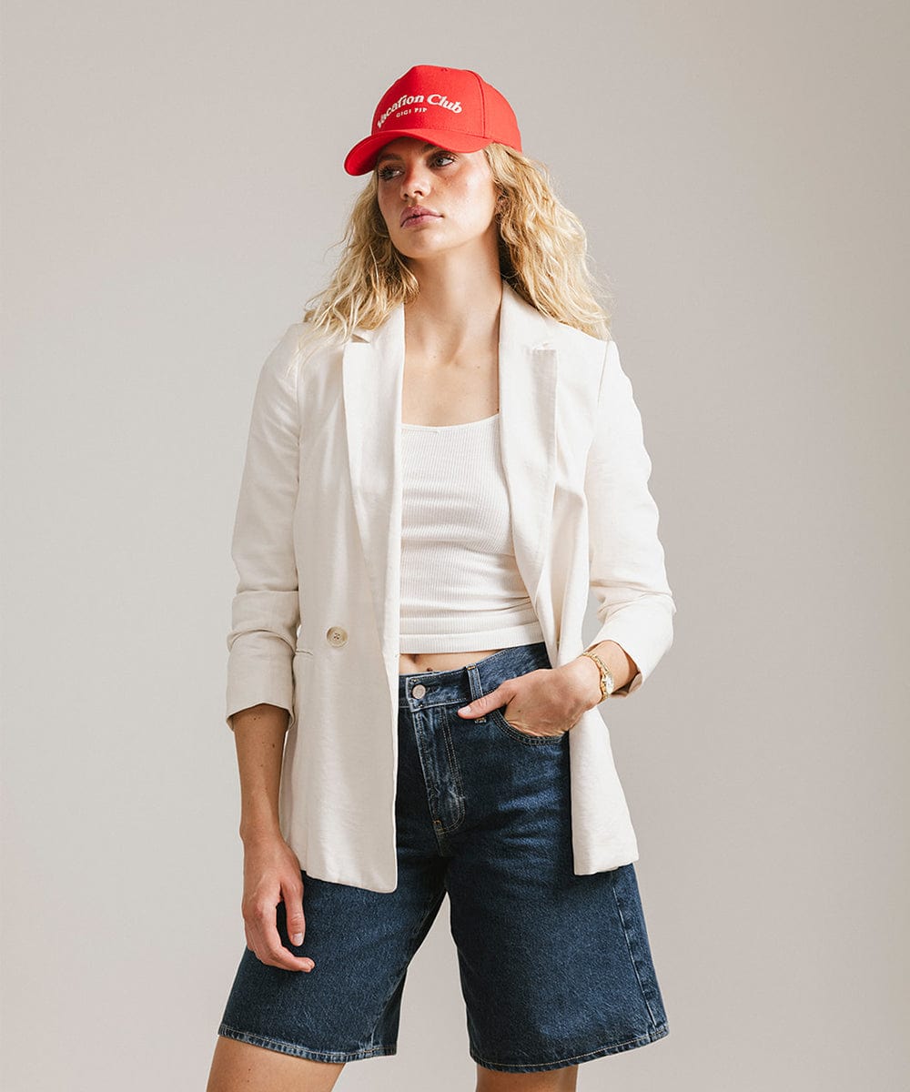 Woman wearing a red cap with 'Vacation Club' text, a white top with a white coat and denim shorts  against a plain background #color_red