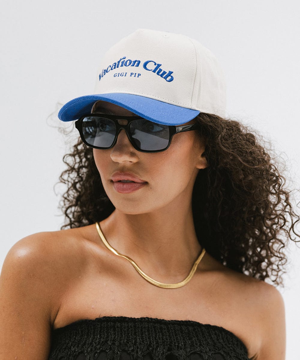 Woman wearing a cream and blue cap with 'Vacation Club' text, sunglasses and a gold chain, posing  against a plain background  #color_cream - blue