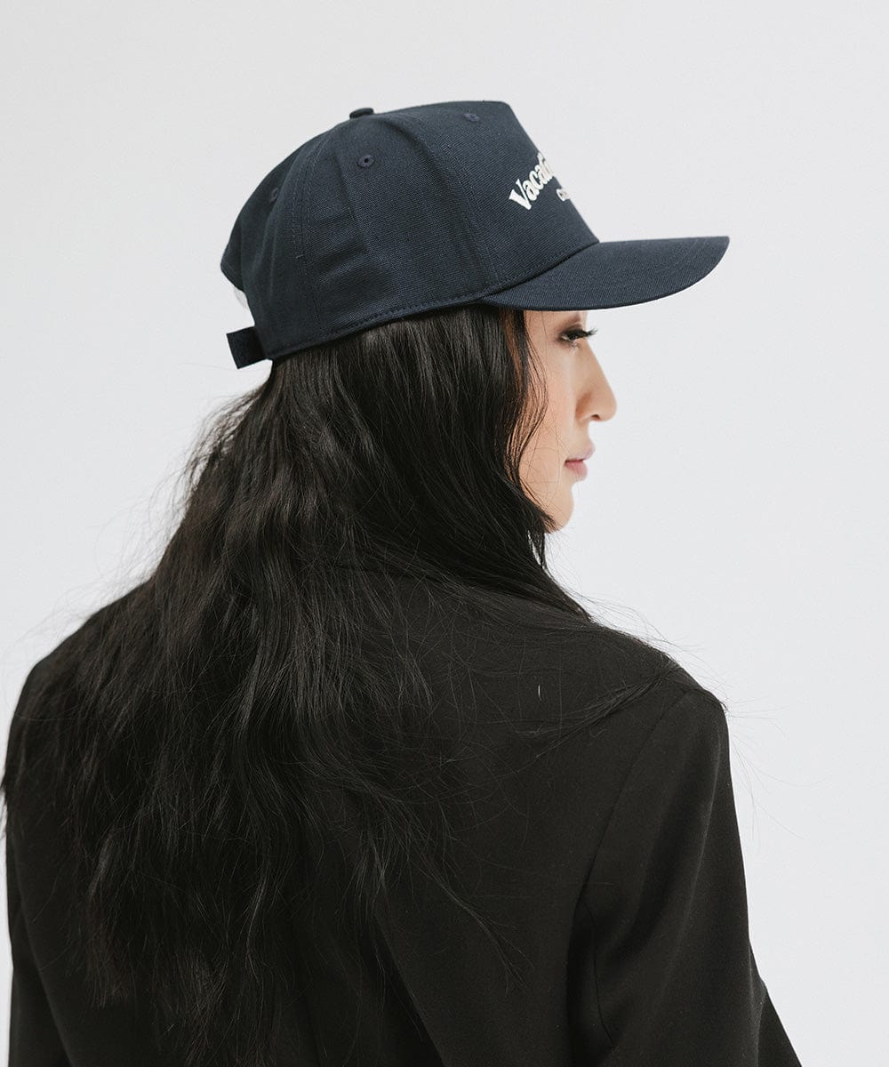 Woman wearing a navy cap and a black blazer, facing sideways against a plain background  #color_navy