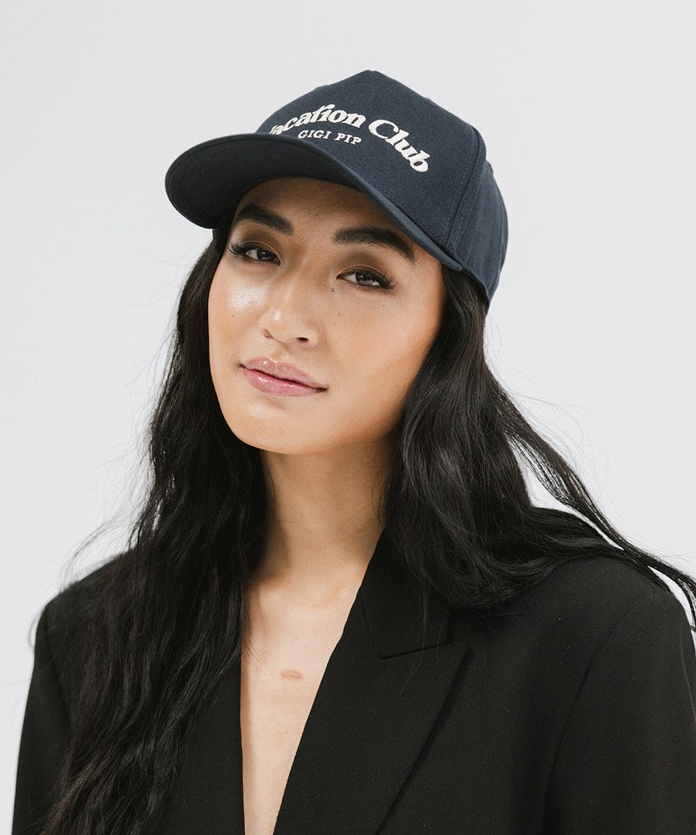 Woman wearing a navy cap with 'Vacation Club' text and a black blazer against a plain background  #color_navy