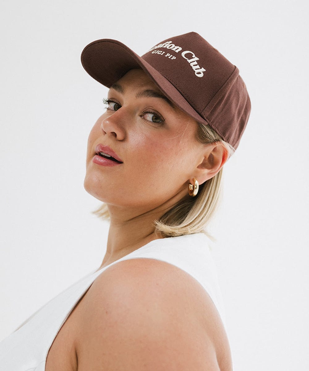 Woman wearing a chocolate brown colored cap with 'Vacation Club' text and a white top, posing sideways  against a plain background #color_chocolate brown
