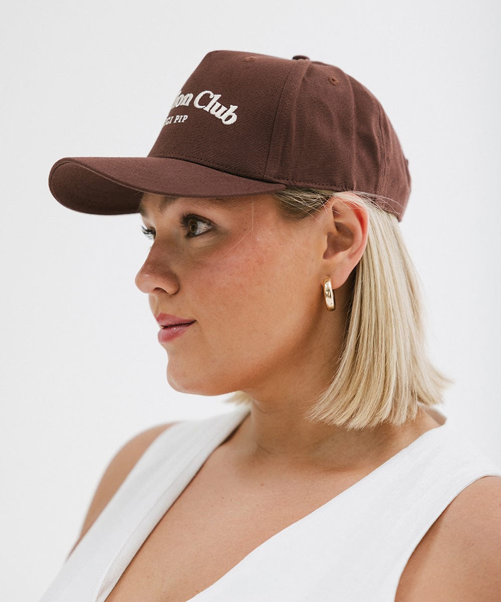 Woman wearing a chocolate brown colored cap with 'Vacation Club' text and a white top, facing sideways  against a plain background #color_chocolate brown