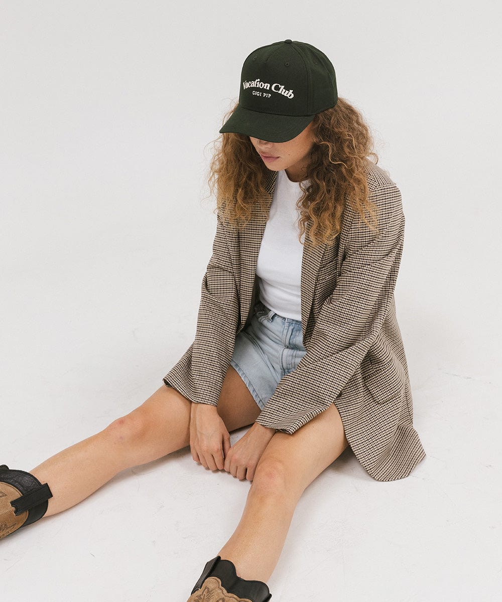 Woman wearing a dark green cap with 'Vacation Club' text, a white top with a coat and denim shorts, sitting  against a plain background  #color_dark green